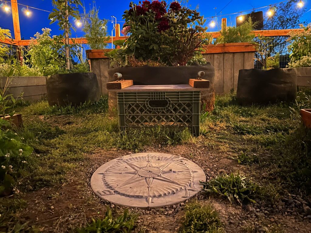 embedded in-ground concrete compass rose facing North with raised beds with dahlias and lit strung lights in the background at twilight