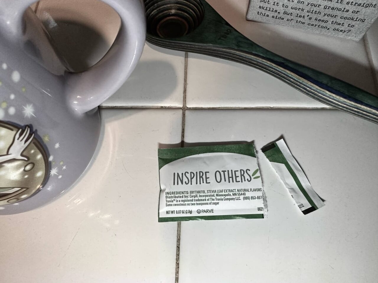 A coffee cup, wooden spoon, oat milk, and sweetener packet that says "inspire others.' Image on About Me page for Sue Evergreen