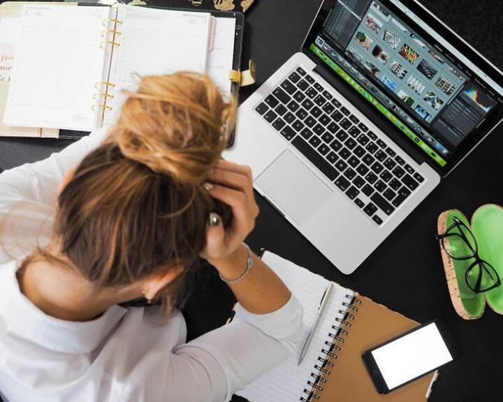 A burnt-out, stressed-out woman in front of her workload of notebooks and a laptop.