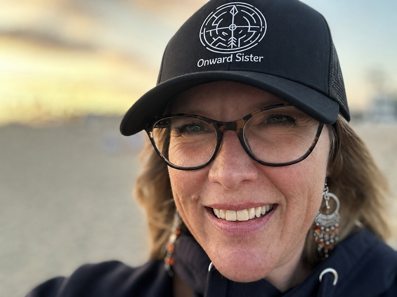 A photo of Sue Evergreen wearing an Onward Sister hat with a sunset in the background. Photo copyrighted by Sue Evergreen.