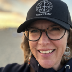 A photo of Sue Evergreen wearing an Onward Sister hat with a sunset in the background. Photo copyrighted by Sue Evergreen.