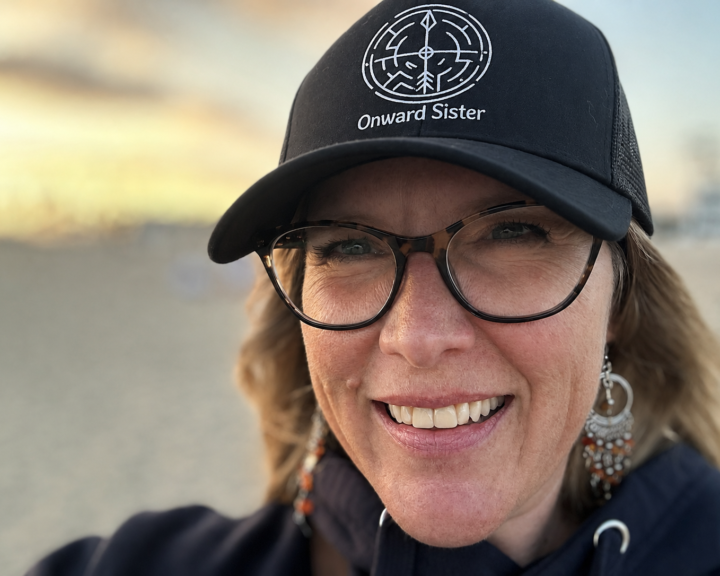 A photo of Sue Evergreen wearing an Onward Sister hat with a sunset in the background. Photo copyrighted by Sue Evergreen.