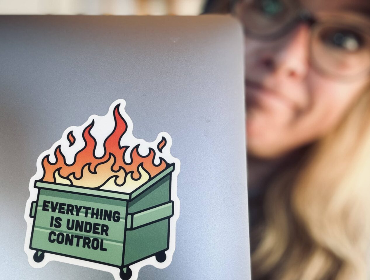 Sue looks out the side of her laptop, which has a dumpster-fire sticker on the front that says, "Everything is under control." Image copyrighted by Dropping Rocks.
