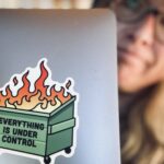 Sue looks out the side of her laptop, which has a dumpster-fire sticker on the front that says, "Everything is under control." Image copyrighted by Dropping Rocks.